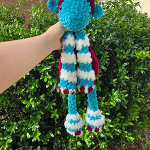 May include: A crocheted plush toy in the shape of a giraffe with a teal blue body, white and red striped legs, and a burgundy head.
