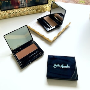 May include: Two black compact cases with mirrors and a brown eyebrow powder inside. The case on the right has the text "Blake Alexander Cosmetics" on the front.