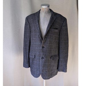 Puede incluir: Una chaqueta de traje gris a cuadros con cierre de dos botones. La chaqueta tiene una sola abertura en la espalda y dos bolsillos de solapa.