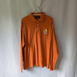 Könnte beinhalten: Orangefarbenes Langarm-Poloshirt mit Bart Simpson-Grafik auf der Vorderseite. Das Shirt hat einen Kragen und drei Knöpfe. Das Simpsons-Logo befindet sich darunter. Das Shirt hängt an einem Holzbügel.