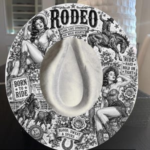 May include: A round, light-coloured hat featuring a detailed black and white graphic print of rodeo scenes, including two cowgirls, cowboys on horses, a bull, roses, and text reading RODEO, LONG LIVE COWBOYS AND WILD HEARTS, BORN TO RIDE, and RIDE HARD HOLD ON TIGHT.