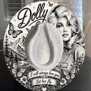 Può includere: Un cappello rotondo di colore chiaro con una stampa grafica in bianco e nero. Il design include un ritratto di Dolly Parton, farfalle, un paesaggio montano e uno stendardo con il testo "I will always love you" e "Let her fly" scritto.