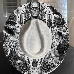 Può includere: Un cappello fedora bianco con una dettagliata stampa grafica in bianco e nero del mostro di Frankenstein e della sua sposa. Il design include rose, un teschio, un cuore, fulmini e striscioni con il testo: WE BELONG DEAD, IT'S ALIVE, TIL DEATH DO US PART e FOREVER MONSTROUS.