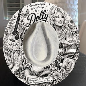 May include: A white felt hat featuring black ink illustrations of Dolly Parton, a guitar, cowboy boots, butterflies, and flowers. Text includes In a world full of ordinary, be a Dolly, Dollywood, Jolene, and Thank you for the music, the laughter and the love.