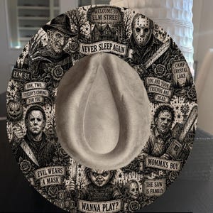 May include: A grey fedora hat featuring a detailed black and white graphic print of iconic horror movie characters and scenes. Text includes Welcome to Elm Street, Never Sleep Again, One, Two, Freddy's Coming For You, You And Your Friends Are Dead, Momma's Boy, Evil Wears A Mask, The Saw Is Family, and Wanna Play?