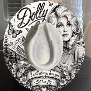 May include: A round, light-coloured hat featuring a detailed black and white illustration of Dolly Parton, butterflies, mountains, and pine trees. The text reads Dolly, I will always love you, Let her fly, and Dolly Tribute at the bottom edge.