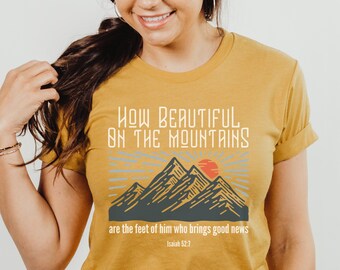 May include: Mustard yellow t-shirt featuring a graphic of mountains and a sunset. The text "HOW BEAUTIFUL ON THE MOUNTAINS are the feet of him who brings good news Isaiah 52:7" is printed on the shirt.