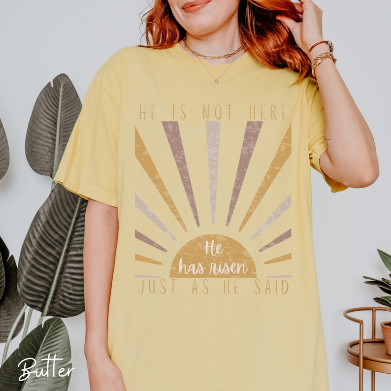 Op de afbeelding: Geel T-shirt met een zonontwerp en de tekst "Hij is niet hier, Hij is opgestaan, Zoals Hij het gezegd heeft."