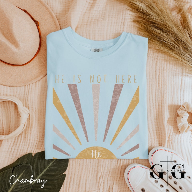 Op de afbeelding: Een lichtblauw t-shirt met een zonsopgang-afbeelding en de tekst "He is not here" in gouden letters. De zon heeft het woord "He" in het midden.