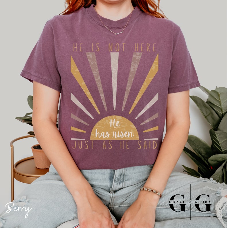 Op de afbeelding: Een paars T-shirt met een zonsopgang-afbeelding en de tekst "Hij is niet hier. Hij is opgestaan. Precies zoals hij het zei."