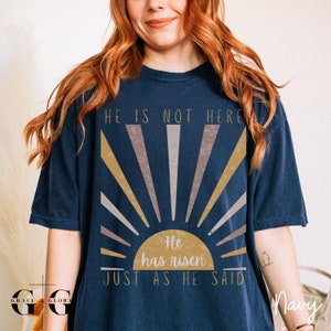 Op de afbeelding: Een marineblauw t-shirt met een afbeelding van een zonsopgang en de tekst "Hij is niet hier, Hij is opgestaan, Zoals Hij het gezegd heeft".