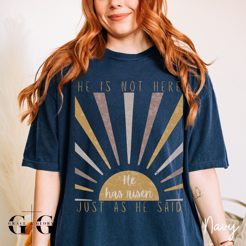 Op de afbeelding: Een marineblauw t-shirt met een afbeelding van een zonsopgang en de tekst "Hij is niet hier, Hij is opgestaan, Zoals Hij het gezegd heeft".