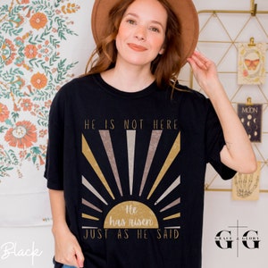 Op de afbeelding: Zwart T-shirt met een zonsopgang ontwerp en de tekst "Hij is niet hier. Hij is opgestaan. Zoals Hij het gezegd heeft."