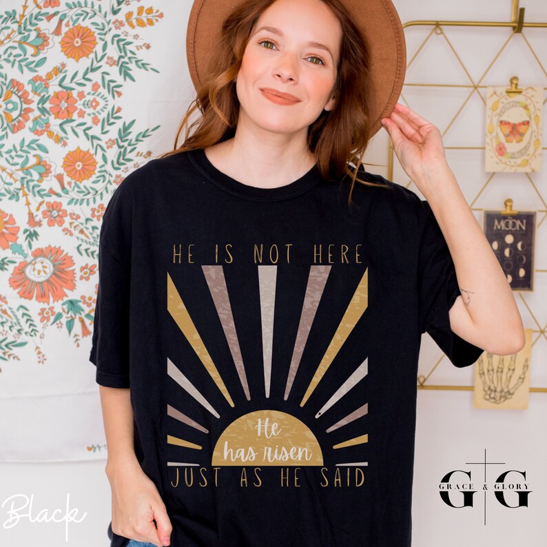 Op de afbeelding: Zwart T-shirt met een zonsopgang ontwerp en de tekst "Hij is niet hier. Hij is opgestaan. Zoals Hij het gezegd heeft."