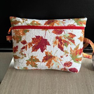 May include: A rectangular pouch with a white background and a pattern of autumn leaves in shades of red, orange, and green. The pouch has a red zipper and a matching strap. The pouch is sitting on a light brown surface.