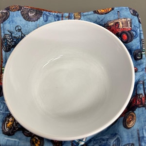 May include: A white ceramic bowl sits on a blue and brown patterned pot holder. The pot holder features a repeating design of vintage tractors in red, blue, and green. The bowl is centered and appears empty.