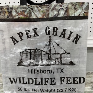 May include: A white tote bag with a camouflage pattern on the top and bottom. The bag has black handles and a black logo that says "APEX GRAIN" with a drawing of grain silos. The text "Hillsboro, TX" and "WILDLIFE FEED" are printed in black below the logo. The text "50 lbs. Net Weight (22.7 KG)" is printed in black at the bottom of the bag.