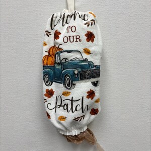 May include: White fabric bag with a blue truck carrying pumpkins, and the words "Welcome to Our Patch". The bag has a loop for hanging and is decorated with fall leaves. A plastic bag is attached to the bottom.