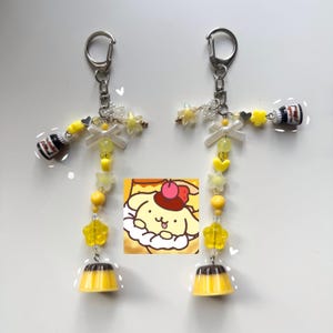 May include: Two keychains with yellow and clear beads, star and heart charms, and miniature pudding and Nutella bottle charms. Each keychain has a silver clasp and a small image of a cartoon character.