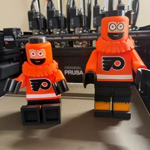 Può includere: Due personaggi in stile Lego arancioni e neri, ciascuno con un logo Flyers sul petto. I personaggi hanno braccia, gambe e mani nere, con colletti e cappelli arancioni. I personaggi sono su una superficie marrone chiaro, con "ORIGINAL PRUSA" visibile sullo sfondo.