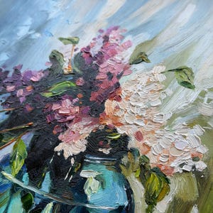 May include: An oil painting featuring a bouquet of purple and white lilac flowers in a blue glass vase. The artwork uses thick, textured brushstrokes to depict the floral clusters, green leaves, and the reflective surface of the vase.