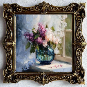 May include: An oil painting in a dark brown and gold ornate frame depicting a bouquet of purple and white lilac flowers in a round blue glass vase. The flowers sit on a white surface near a window with soft light and visible brushstrokes.