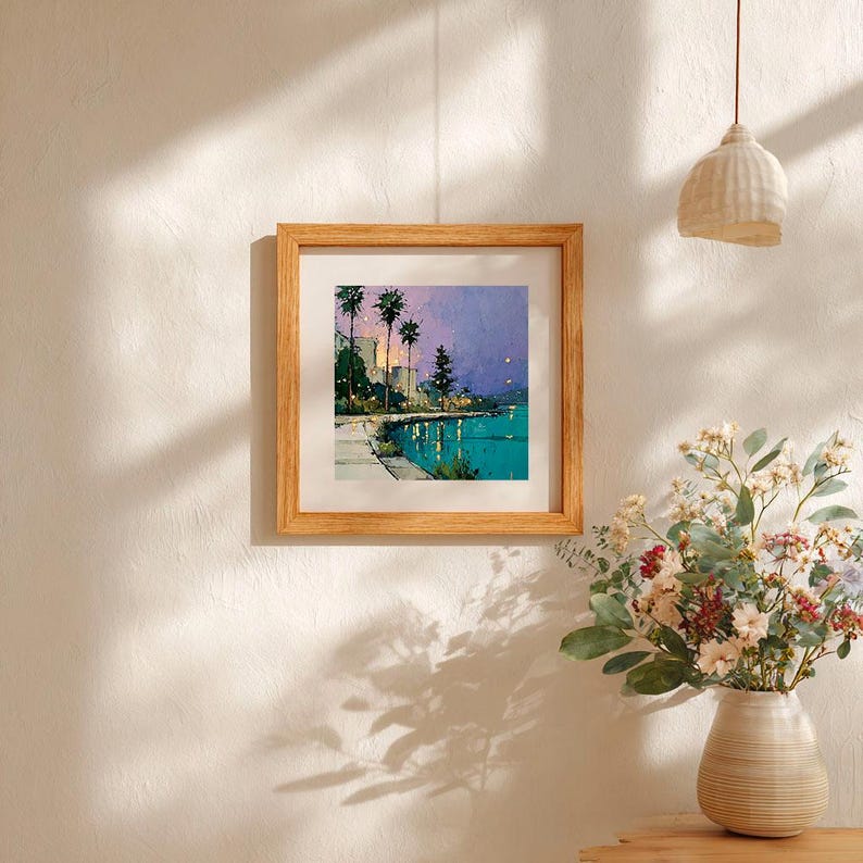 May include: A framed art print depicting a coastal scene with palm trees, buildings, and a body of water. The artwork features a color palette of teal, purple, and yellow, and is encased in a light brown wooden frame. The print is displayed on a white wall.