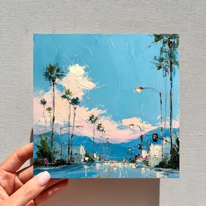 May include: An oil painting on canvas depicting a street scene with tall palm trees under a bright blue sky. The artwork features thick, textured brushstrokes in shades of blue, white, and soft pink, capturing a sunny day with light clouds.
