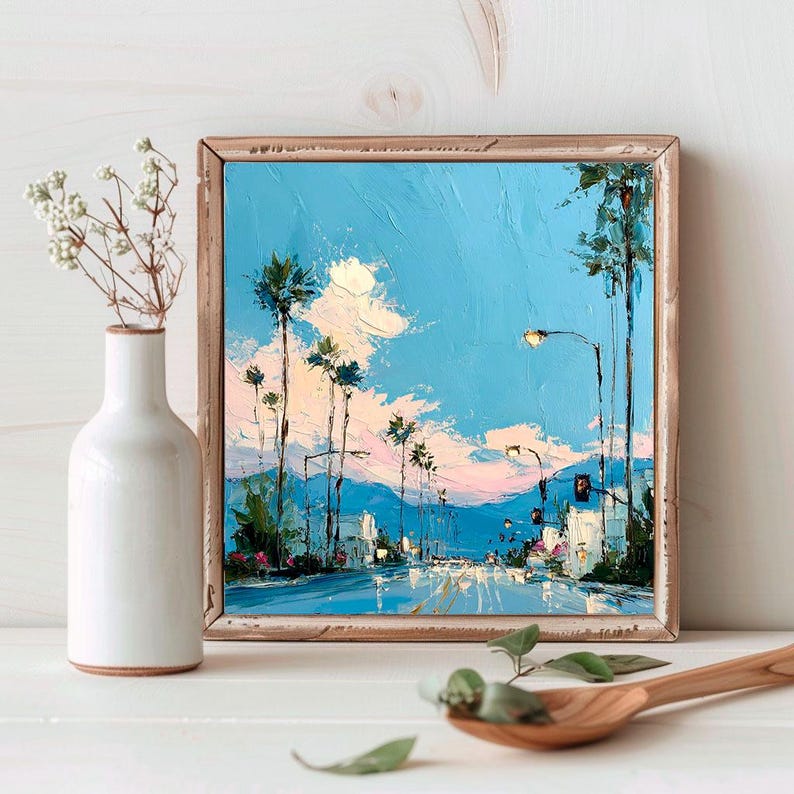 May include: A square oil painting in a wooden frame depicting a street scene with tall palm trees under a bright blue sky with soft pink and white clouds. The artwork features thick, textured brushstrokes and warm light from street lamps.