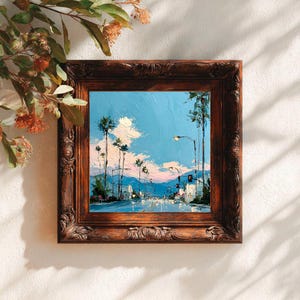 May include: A square oil painting in a dark brown ornate wooden frame depicting a street scene with tall palm trees against a bright blue sky with pink and white clouds. The artwork features thick, textured brushstrokes and a road perspective.