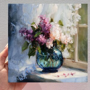 May include: An oil painting on canvas depicting a bouquet of purple and white lilac flowers arranged in a round, translucent blue glass vase. The flowers are positioned near a window with light filtering through, casting soft shadows and highlights.