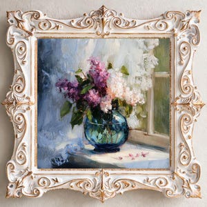 May include: An oil painting in a white and gold ornate frame depicting a bouquet of purple and white lilacs in a round blue glass vase. The flowers sit on a surface near a window with soft light, featuring visible brushstrokes and textures.