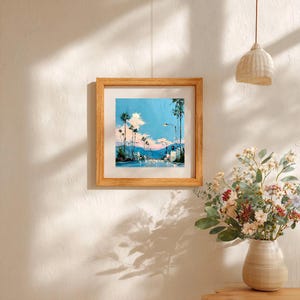 May include: A square framed painting depicting a street scene with tall palm trees under a bright blue sky with white clouds. The artwork features a light wooden frame and is displayed on a wall, showcasing a vibrant landscape with soft pink hues.