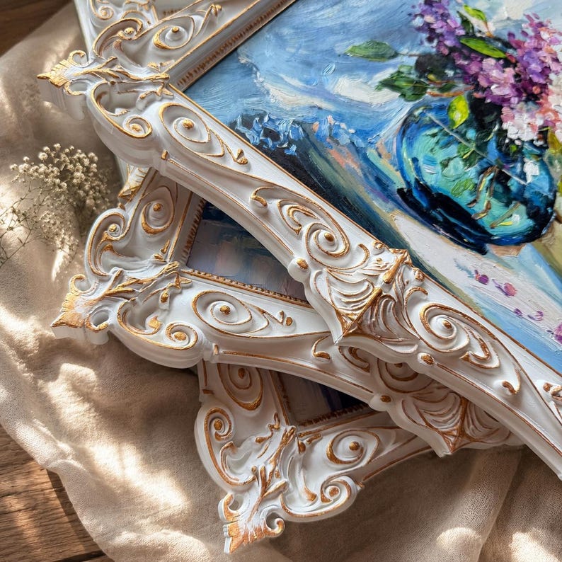 May include: An oil painting in a white and gold ornate frame featuring a blue vase filled with purple lilac flowers. The painting uses thick, textured brushstrokes to depict the floral arrangement against a light blue background with visible paint.