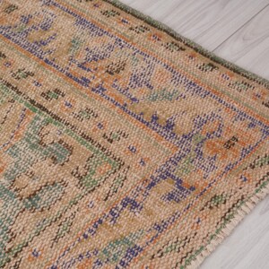 Pu&ograve; includere: Primo piano di una sezione di un tappeto vintage dall'aspetto usurato e sbiadito. Il tappeto presenta uno sfondo beige con motivi intricati nei toni del blu, dell'arancione e del verde. I bordi del tappeto sono visibili, mostrando una trama usurata.