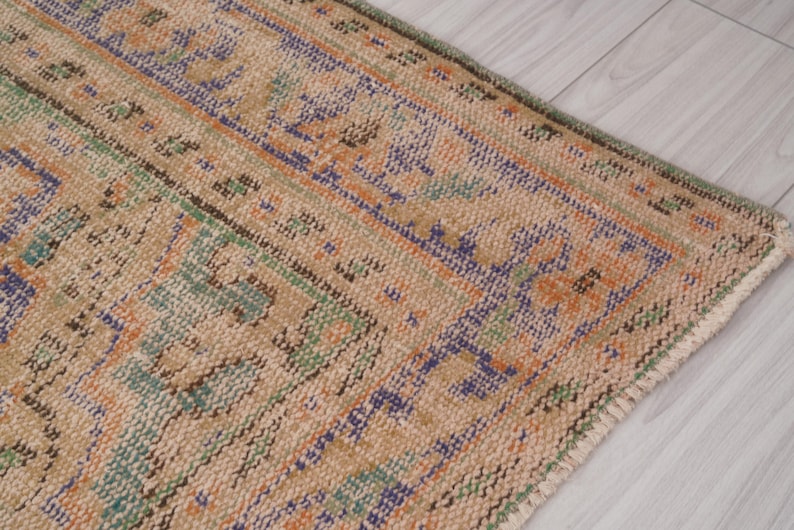 Pu&ograve; includere: Primo piano di una sezione di un tappeto vintage dall'aspetto usurato e sbiadito. Il tappeto presenta uno sfondo beige con motivi intricati nei toni del blu, dell'arancione e del verde. I bordi del tappeto sono visibili, mostrando una trama usurata.