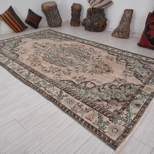 May include: A rectangular, vintage-style rug with a central medallion design. The rug features a beige background with floral patterns in shades of green, brown, and purple. The border is a darker brown with intricate detailing. The rug is approximately 183 x 274 cm.