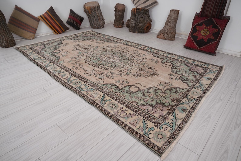 May include: A rectangular, vintage-style rug with a central medallion design. The rug features a beige background with floral patterns in shades of green, brown, and purple. The border is a darker brown with intricate detailing. The rug is approximately 183 x 274 cm.