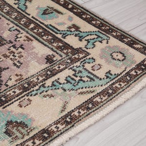 May include: Detail of a vintage rug with a distressed design. The rug has a cream-coloured background with a brown and teal border. The central design features muted shades of green and purple.