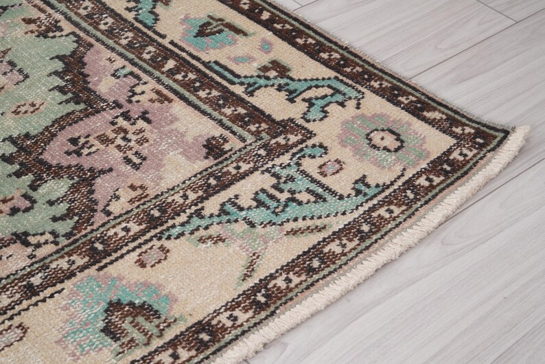 May include: Detail of a vintage rug with a distressed design. The rug has a cream-coloured background with a brown and teal border. The central design features muted shades of green and purple.