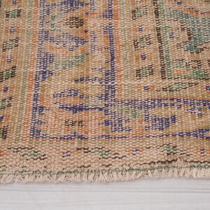 Pu&ograve; includere: Primo piano di una sezione di un tappeto vintage dall'aspetto usurato e sbiadito. Il tappeto presenta uno sfondo beige con motivi intricati in blu, arancione e verde. I bordi hanno un dettaglio a frange.
