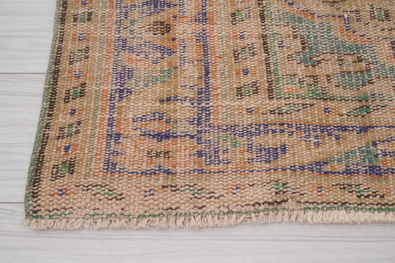 Pu&ograve; includere: Primo piano di una sezione di un tappeto vintage dall'aspetto usurato e sbiadito. Il tappeto presenta uno sfondo beige con motivi intricati in blu, arancione e verde. I bordi hanno un dettaglio a frange.