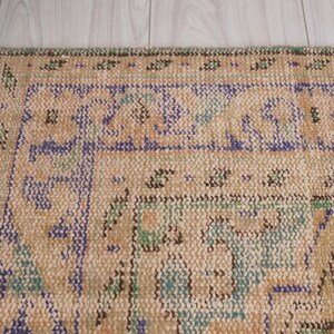 Pu&ograve; includere: Primo piano di una sezione di un tappeto vintage con sfondo beige e motivi intricati in blu, verde e marrone. Il tappeto ha un bordo sfrangiato e un aspetto usurato, che suggerisce et&agrave; e utilizzo. Il design presenta motivi geometrici e floreali.