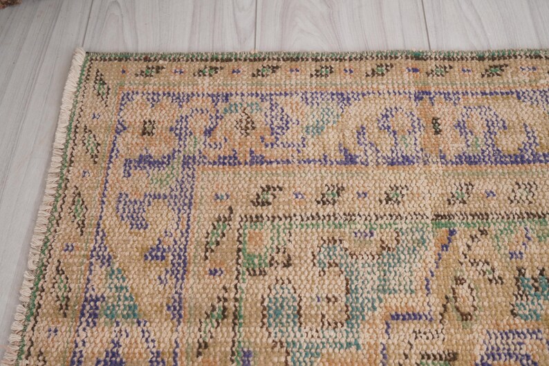 Pu&ograve; includere: Primo piano di una sezione di un tappeto vintage con sfondo beige e motivi intricati in blu, verde e marrone. Il tappeto ha un bordo sfrangiato e un aspetto usurato, che suggerisce et&agrave; e utilizzo. Il design presenta motivi geometrici e floreali.
