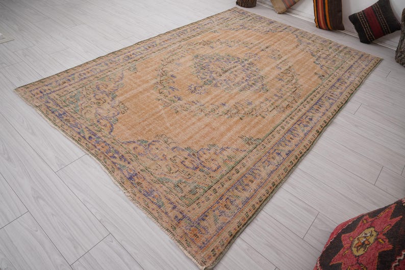 Pu&ograve; includere: Un tappeto rettangolare dall'aspetto invecchiato, con una palette di colori pesca e beige sbiaditi. Il tappeto presenta un disegno floreale centrale con accenti di blu, verde e viola. Il bordo ha un motivo dettagliato. Il tappeto &egrave; posato su un pavimento in legno chiaro.