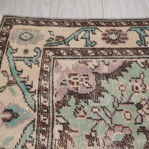 May include: Detail of a vintage rug with a faded design. The rug has a cream-coloured central area with a floral pattern in green, purple, and brown. The border is brown with teal accents. Fringes are visible along the edge.