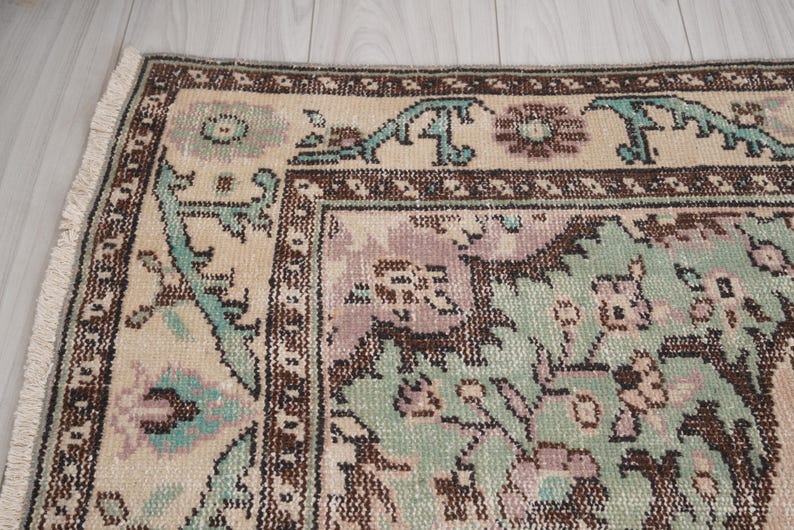 May include: Detail of a vintage rug with a faded design. The rug has a cream-coloured central area with a floral pattern in green, purple, and brown. The border is brown with teal accents. Fringes are visible along the edge.