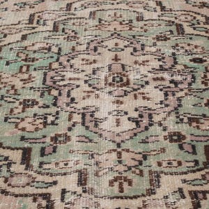 May include: Close-up of a vintage rug with a detailed, symmetrical pattern. The rug features a central medallion design in shades of green, brown, and beige, with intricate floral and geometric motifs. The rug has a distressed, antique appearance.