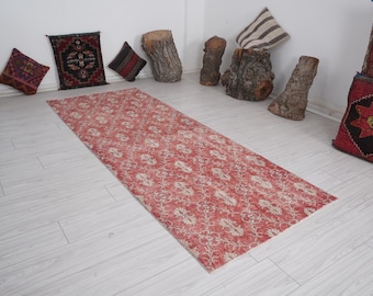 May include: A long, rectangular, red and cream-colored rug with a floral pattern. The rug is placed on a light-colored wooden floor. Decorative pillows and wooden stumps are in the background.
