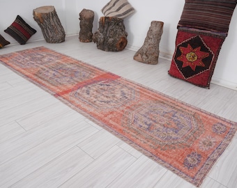 Op de afbeelding: Een lange, rechthoekige, versleten vloerkleed met een vervaagde, vintage uitstraling. Het vloerkleed heeft een centraal ontwerp in tinten oranje, rood en paars, met een patroonrand. Het vloerkleed ligt op een lichtgekleurde houten vloer.